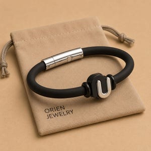 May include: A black silicone bracelet with a silver clasp and a silver "U" shaped accent. The bracelet is displayed on a beige pouch with the text "ORIEN JEWELRY".