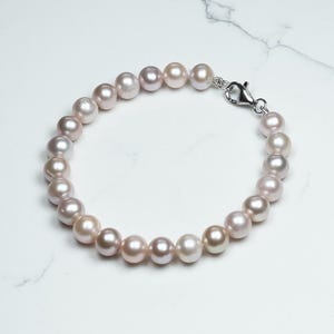 May include: A delicate bracelet featuring a strand of lustrous, light pink pearls. The pearls are round and evenly sized, strung together with a silver-colored clasp. The bracelet is displayed on a white surface, highlighting the pearls' soft color.