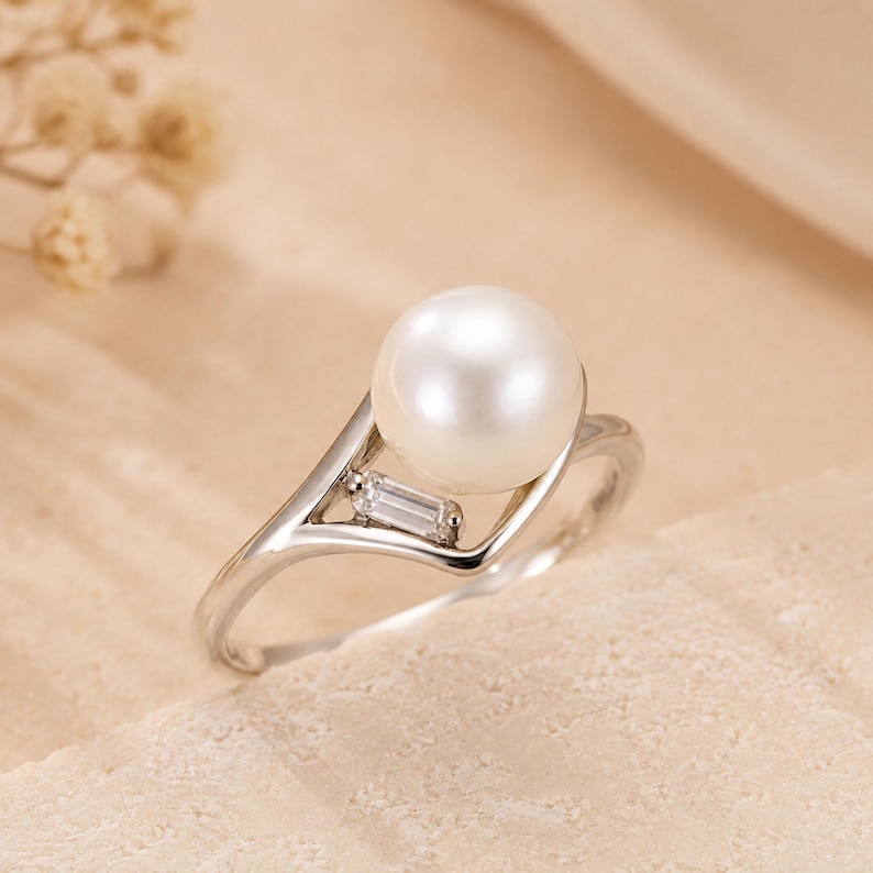 May include: A silver-toned ring featuring a single round white pearl set at the top, accented by a small, rectangular baguette-cut clear stone positioned on the side of the band, creating a sleek and polished look for this jewelry piece.