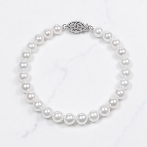 May include: A single strand of round, white, lustrous pearls arranged in a circular formation. The necklace features a decorative, oval-shaped silver-toned metal clasp with an intricate, filigree-style cutout pattern at the top center.