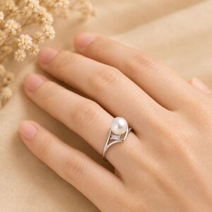 May include: A silver-toned ring featuring a single round white pearl and a small rectangular baguette-cut clear stone set into a split shank band. The metal band curves around the pearl, creating an open design that highlights the gemstone.