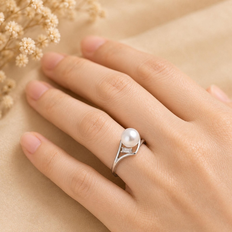 May include: A silver-toned ring featuring a single round white pearl and a small rectangular baguette-cut clear stone set into a split shank band. The metal band curves around the pearl, creating an open design that highlights the gemstone.
