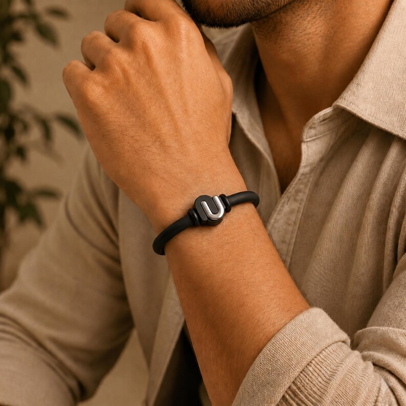 May include: A black bracelet with a silver-colored "U" shaped charm. The bracelet is made of a flexible material and is worn on the wrist. The charm is centered on the bracelet.