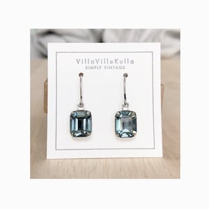 May include: A pair of silver dangle earrings with dark gray square-shaped crystal stones. The earrings are displayed on a white card with the text "VillaVilleKulla Simply Vintage".