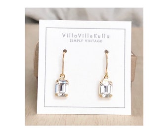 May include: A pair of gold dangle earrings with clear emerald-cut crystal stones. The earrings are displayed on a white card with the text "VillaVilleKulla Simply Vintage".