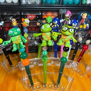 May include: A collection of decorative pens featuring various cartoon character toppers. The pens have black or red barrels and are displayed in clear plastic stands. The toppers include characters like turtles, Minions, and Sonic the Hedgehog.