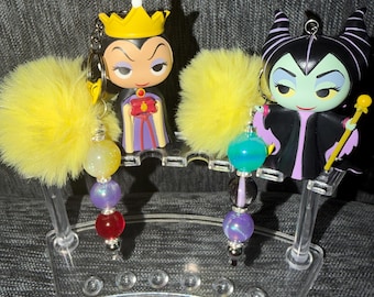 May include: Two character keychains featuring the Evil Queen and Maleficent, each attached to a yellow faux fur pom-pom and a strand of three colorful beads. The keychains are displayed on a clear, curved acrylic stand with multiple circular slots.