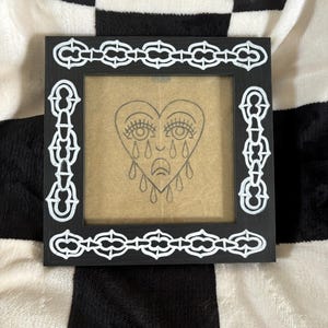 Puede incluir: Marco de imagen negro con un borde de cadena blanca. El marco contiene un dibujo de un corazón con una cara que está llorando.