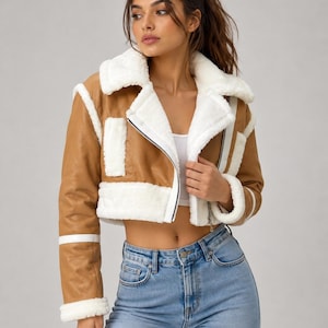 May include: A cropped tan jacket with white faux shearling trim on the collar, cuffs, and hem. The jacket features a front zipper closure, long sleeves with white horizontal stripes, and a white cropped top worn underneath with light blue jeans.