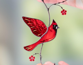 May include: A stained glass cardinal bird with outstretched wings, perched on a metal branch with red flowers. The bird is a vibrant red with black accents, and the metal has a dark finish. The piece is held up by a loop at the top.