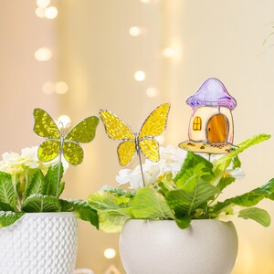 Può includere: Due piante in vaso con decorativi picchetti da giardino. Un vaso ha due picchetti a farfalla gialli, e l'altro un picchetto a forma di casetta delle fate. I vasi sono bianchi e beige, e le piante hanno foglie verdi e fiori bianchi.