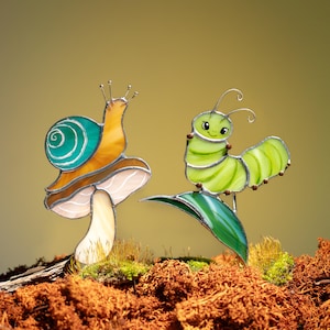 May include: Two stained glass garden ornaments: a snail on a mushroom and a caterpillar on a leaf. The snail has a teal shell and an orange body. The caterpillar is green. The mushroom is brown and white. The leaf is green.