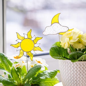 Könnte beinhalten: Zwei Topfpflanzen mit gelben Blüten und dekorativen Gartensteckern in Form von Sonne und Mond. Der Sonnenstecker ist gelb mit schwarzer Umrandung, der Mond gelb mit einer weißen Wolke. Die Töpfe sind weiß und stehen auf einer Holzoberfläche.