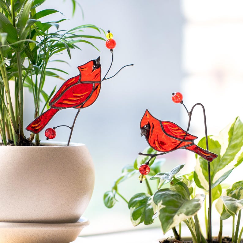 May include: Two red cardinal bird garden stakes with black metal stems and berry accents. The birds are perched on branches among green plants in a beige pot, adding a touch of nature to indoor or outdoor decor.