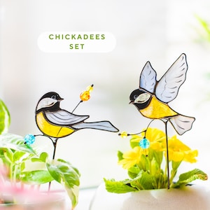May include: A pair of decorative chickadee bird stakes. The birds are made of stained glass in yellow, black, and grey, with beaded details. The text "CHICKADEES SET" is above the birds. The stakes are placed in potted plants and flowers.