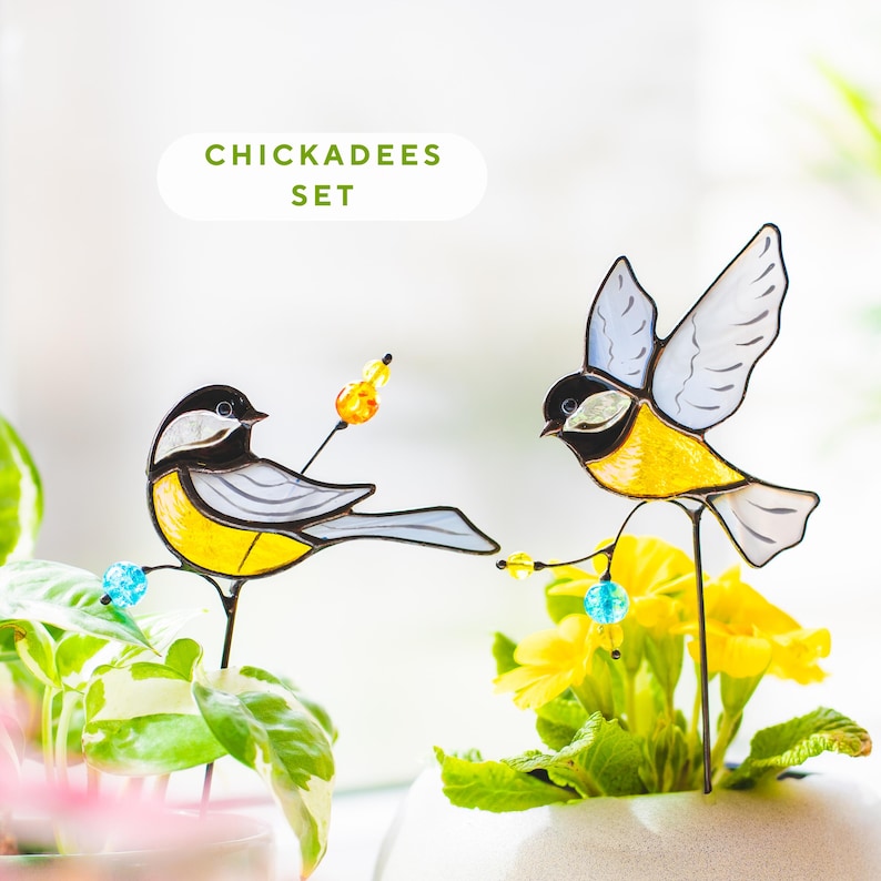 May include: A pair of decorative chickadee bird stakes. The birds are made of stained glass in yellow, black, and grey, with beaded details. The text "CHICKADEES SET" is above the birds. The stakes are placed in potted plants and flowers.