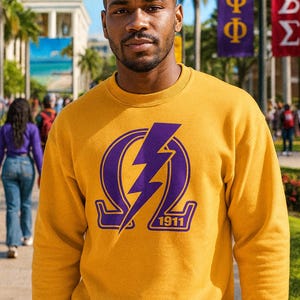 May include: Mustard yellow sweatshirt with a purple lightning bolt and Greek letters design. The sweatshirt has a crew neck and long sleeves. The design includes the year 1911. The background shows a college campus with banners.