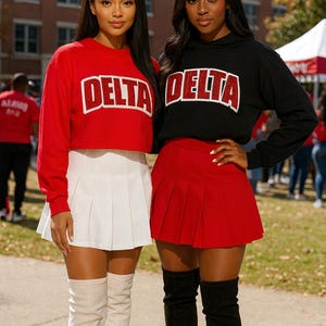 May include: Two women wearing matching outfits. One wears a red cropped sweatshirt with "DELTA" in white block letters, a white pleated skirt, and beige thigh-high boots. The other wears a black hoodie with "DELTA" and a red pleated skirt, with black thigh-high boots.