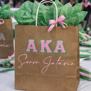 Puede incluir: Bolsa de regalo de papel marrón con asas blancas, llena de papel de seda verde. La bolsa tiene las letras "AKA" en rosa y verde, y el nombre "Soror Jataiva" en escritura rosa. Un lazo rosa adorna la bolsa.