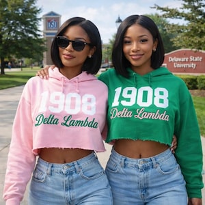 May include: Two women wearing matching cropped hoodies and light wash jeans. One hoodie is pink, the other is green. Both hoodies have the text "1908 Delta Lambda" in white and green script. Sunglasses are worn by one woman.