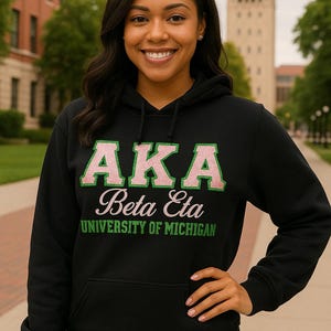May include: Black hooded sweatshirt with the letters "AKA" in green and pink, with "Beta Eta" and "University of Michigan" printed below. The sweatshirt has a front pocket and is worn by a person.