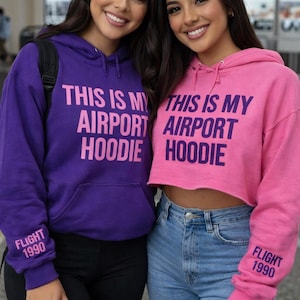 May include: Two people wearing purple and pink hoodies with the text "THIS IS MY AIRPORT HOODIE" printed on the front. The purple hoodie has "FLIGHT 1990" printed on the sleeve. The pink hoodie is cropped and paired with blue jeans.