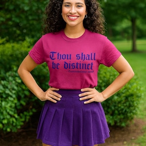 Peut inclure: Une femme porte un t-shirt magenta avec l'inscription "Thou shall be distinct" en bleu, une jupe plissée violette et des chaussettes hautes assorties. L'arrière-plan est un environnement verdoyant.