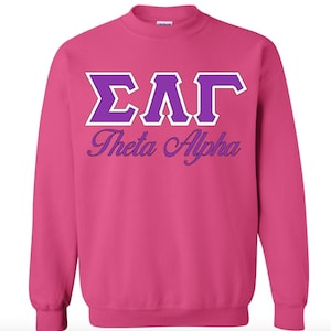 Peut inclure: Sweat-shirt rose avec des lettres blanches et violettes qui disent "ΣΑΓ Theta Alpha".
