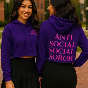 Peut inclure: Deux femmes portant des sweats à capuche violets courts avec le texte "ANTI SOCIAL SOCIAL SOROR" en rose au dos. Les sweats à capuche ont des cordons de serrage et sont associés à des jupes noires. L'image est prise à l'extérieur.