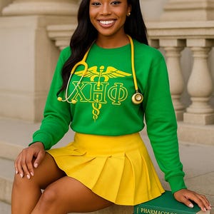 Peut inclure: Une femme porte une coiffe de graduation, un sweat-shirt vert avec des lettres grecques jaunes et une jupe plissée jaune. Un stéthoscope jaune est autour de son cou. Elle est assise sur des marches avec des livres intitulés "Pharmacologie", "Recherche en soins infirmiers" et "Maternité et santé des femmes".