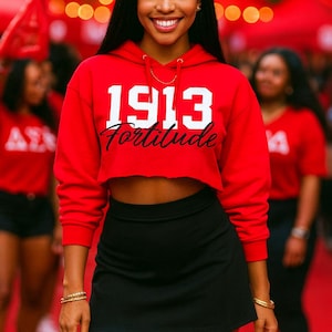 May include: A red cropped hoodie with the text "1913 Fortitude" in white and black, paired with a black mini skirt. The image features a woman smiling, with other people in the background.