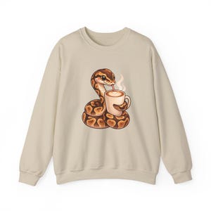May include: A beige crewneck sweatshirt featuring a graphic illustration of a brown and tan patterned snake coiled around a white mug of steaming coffee with latte art. The snake has large eyes and is drinking from the mug with a thin straw.