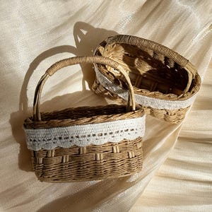 May include: Two small, woven wicker baskets with handles and white lace trim. One basket is upright, the other is on its side. The baskets are brown and beige, and sit on a cream-coloured, textured fabric.