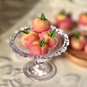 May include: A glass pedestal cake stand with a clear glass base and a scalloped edge. The stand is filled with five miniature peach-colored fruit, each with a green leaf on top.