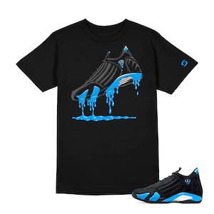 May include: Black t-shirt featuring a graphic of a black and blue sneaker with dripping blue paint. A matching black and blue sneaker is in the lower right corner. The shirt has a small blue design on the right sleeve.