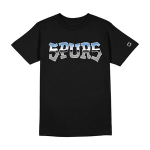May include: A black short-sleeved t-shirt featuring the word SPURS printed across the chest in a metallic-style font with a blue and silver gradient. A small silver logo is visible on the left sleeve near the shoulder seam.
