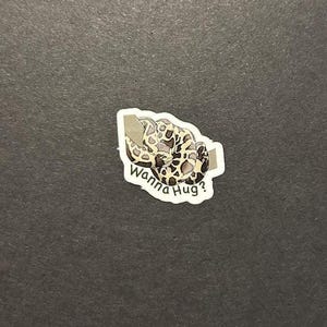 May include: A sticker featuring a coiled snake with a brown and cream pattern. The snake is wrapped around a gray rectangle. The text "Wanna Hug?" is written in a playful font below the snake.