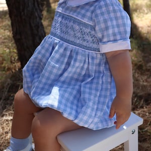 Puede incluir: Un bebé con un vestido smockado de cuadros azules y blancos con cuello blanco. El vestido tiene un corpiño smockado y una falda fruncida. El bebé está sentado en un taburete blanco.