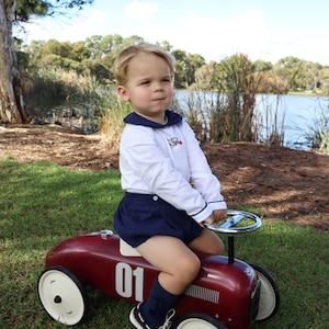 May include: A young child in a white shirt with a navy collar and shorts sits on a vintage red toy car with white wheels. The car has the number "01" on the side. The child wears navy socks and black and white shoes. The scene is outdoors.