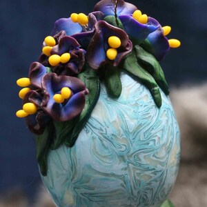 May include: An egg-shaped ornament with a marbled blue and white design. It features purple flowers with yellow centres and green leaves. A thin gold wire is attached for hanging, suitable for decoration.