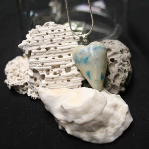 May include: A white heart-shaped pendant with blue swirls, hanging from a silver chain. The pendant is resting on a white seashell.