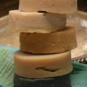 Puede incluir: Una pila de cinco barras de jabón redondas, con la barra superior de color rosa claro, la segunda de color beige claro, la tercera de color marrón claro, la cuarta de color rosa claro y la barra inferior de color gris oscuro.