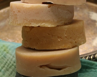 May include: A stack of five round bars of soap, with the top bar being a light pink color, the second bar being a light beige color, the third bar being a light brown color, the fourth bar being a light pink color, and the bottom bar being a dark gray color.