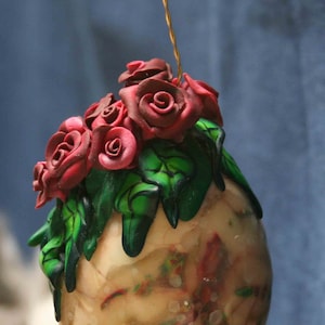 以下が含まれることがあります： 花柄のデザインが施された卵型のオーナメント。オーナメントの上部には赤いバラと緑の葉が飾られています。卵は赤と緑のアクセントで大理石模様になっています。吊り下げ用の金色のワイヤーが付いています。