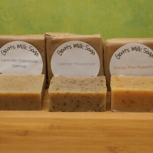 May include: A selection of goat's milk soaps, wrapped in brown paper with white labels. Flavours include Oatmeal Nutmeg Honey, Lavender Chamomile Oatmeal, Lavender Peppermint, Orange Pine Peppermint, and Lavender Pine. Soaps are displayed on a wooden tray.