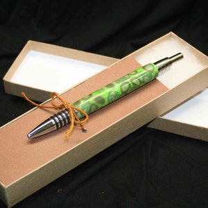 Könnte beinhalten: Ein grüner und silberner Stift mit einem Wirbelmuster wird in einer braunen Schachtel mit weißem Innenraum gezeigt. Der Stift ist mit einer braunen Schnur gebunden.