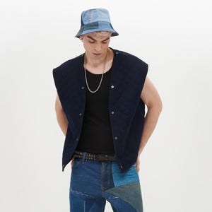 Puede incluir: Un chaleco acolchado azul marino con cierre de presi&oacute;n, usado sobre una camiseta negra de tirantes. La persona lleva un sombrero de pescador de mezclilla azul con parches y pantalones de mezclilla azul con parches y un cintur&oacute;n negro con tachuelas.