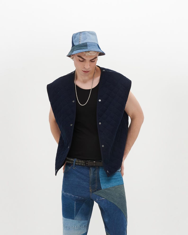 Puede incluir: Un chaleco acolchado azul marino con cierre de presi&oacute;n, usado sobre una camiseta negra de tirantes. La persona lleva un sombrero de pescador de mezclilla azul con parches y pantalones de mezclilla azul con parches y un cintur&oacute;n negro con tachuelas.