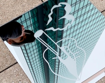 May include: A square mirror with a white graphic of a hand holding a cigarette, with smoke rising. The mirror reflects a person smoking a cigarette and a tall building with many windows. The mirror is on a gray surface.