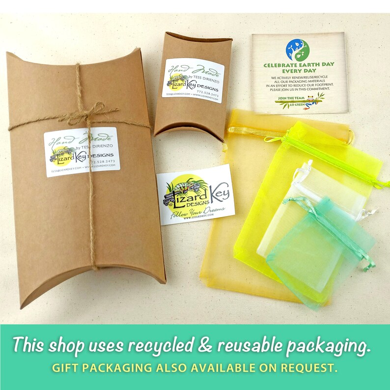 May include: A close-up of a brown cardboard box with a white label that reads "Hand Made by Tess Dirienzo Lizard Key Designs". The box is tied with twine and is sitting on a white surface. There are also two yellow and green mesh bags, a small white card with the text "Lizard Key Designs Follow Your Dreams", and a small card with the text "Celebrate Earth Day Every Day" in the image.
